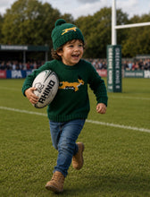 Child wearing green and gold hand-knitted beanie and jersey with animal design, playing rugby outdoors