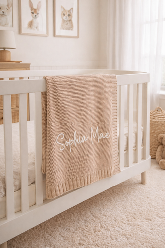 Personalised cotton knit baby blanket in camel, draped over a nursery crib